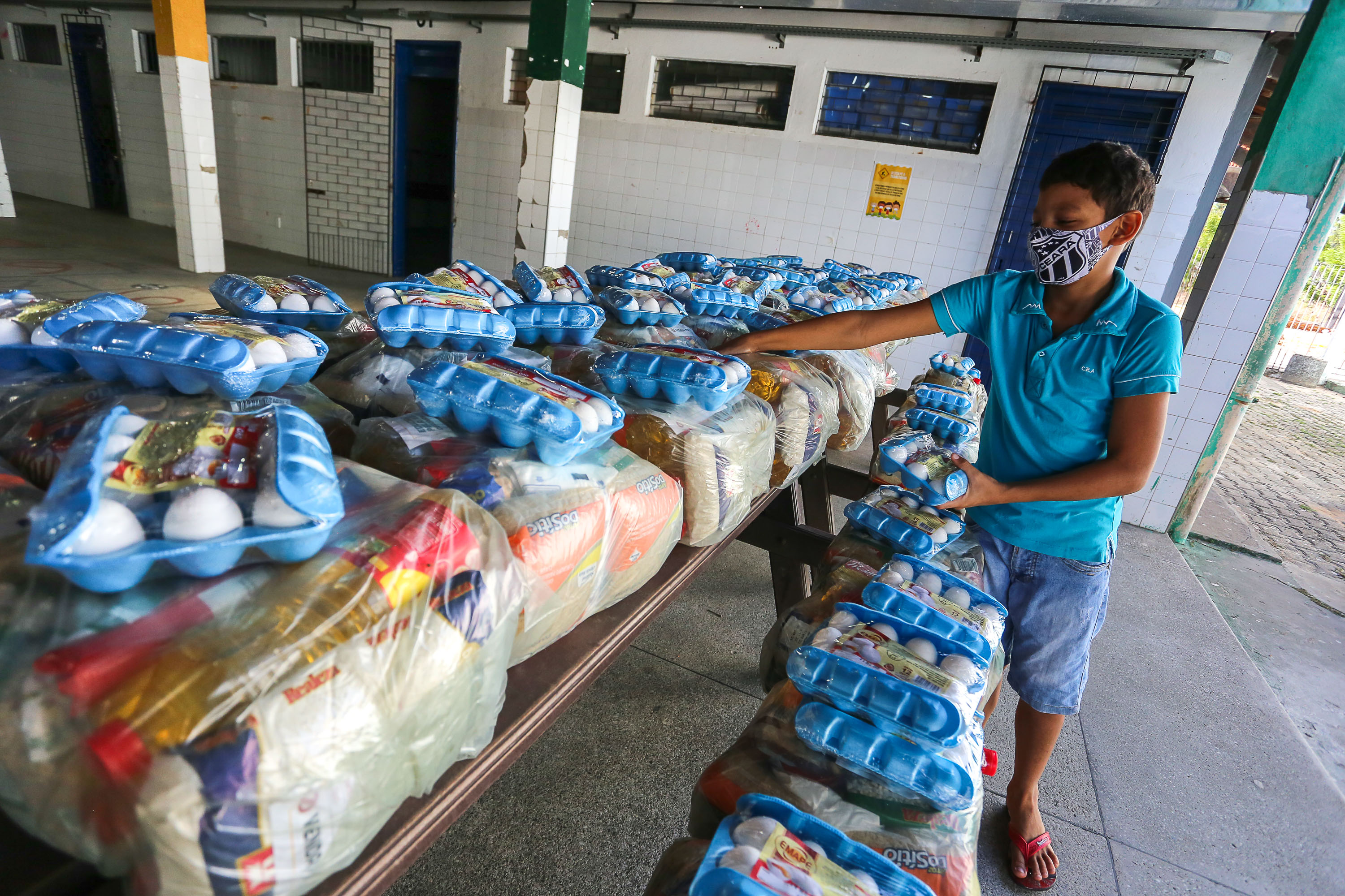 garoto usando mácara pegando pegando kits alimentares em pilha com vários kits